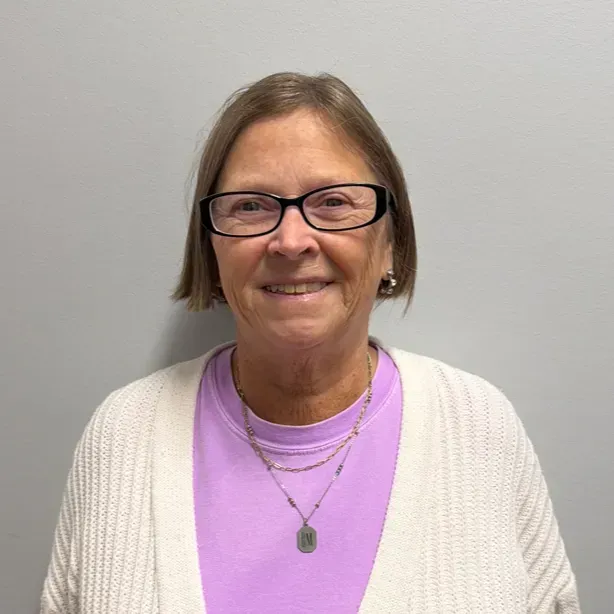 Woman wearing glasses, a light purple shirt, and a cream cardigan smiles in front of a gray wall.