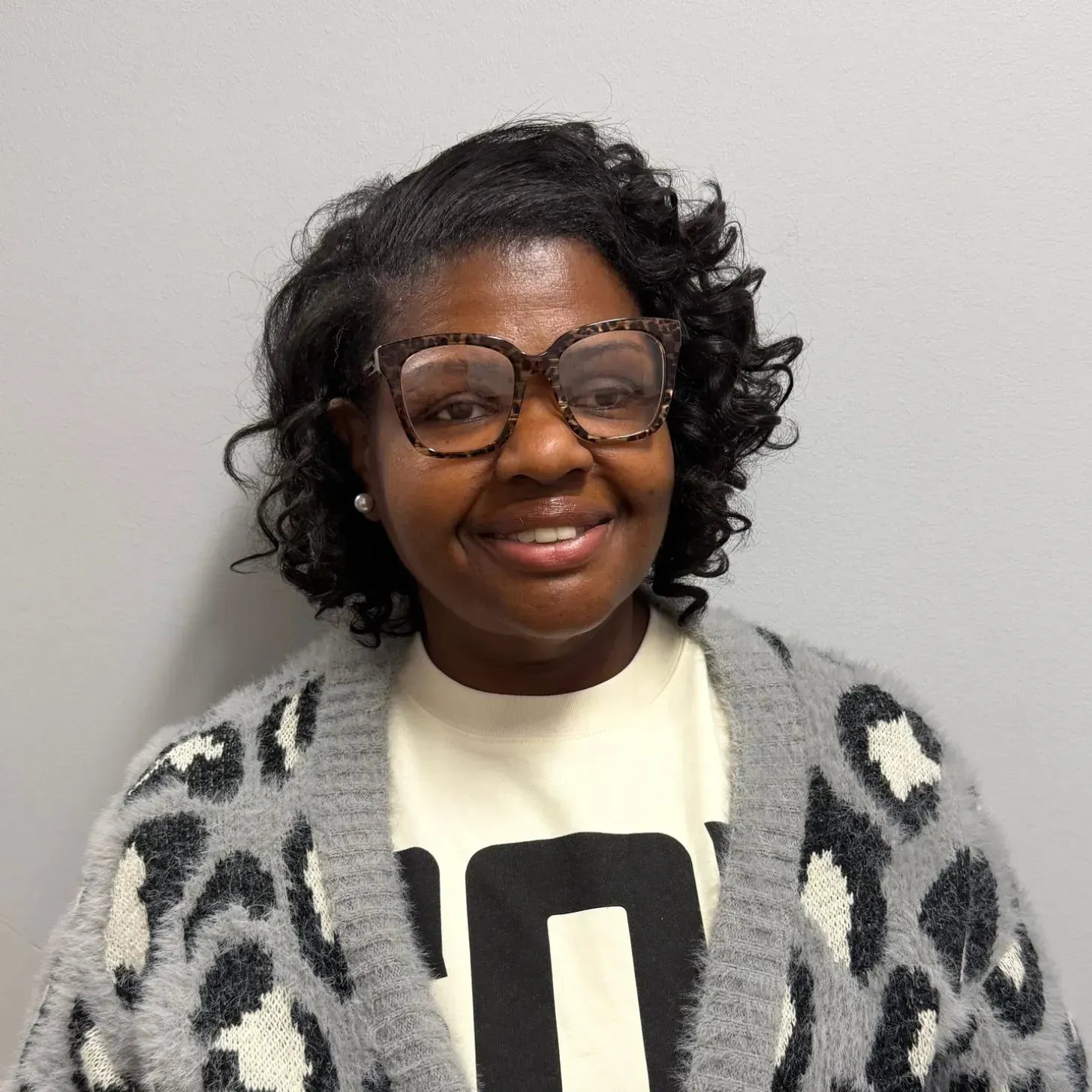 Woman with glasses smiles, wearing a cream shirt and grey leopard print cardigan in front of a grey wall.