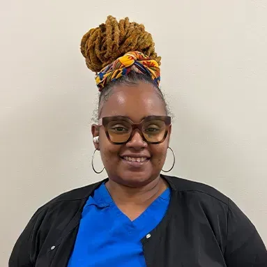Woman with glasses and wrapped hair smiles. She wears a blue top and black jacket.