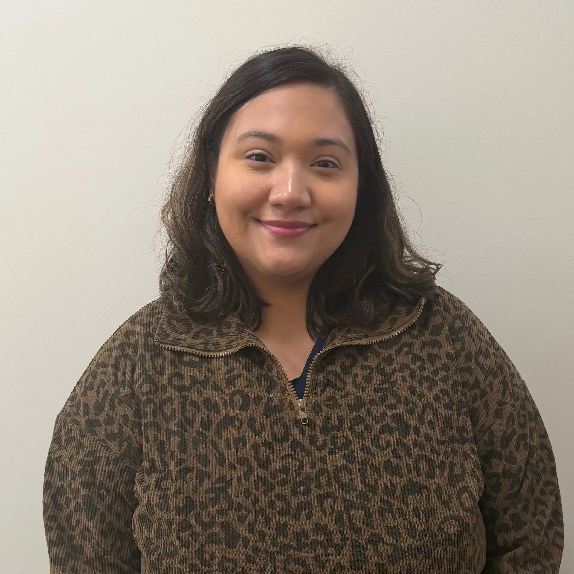 Woman in a brown and black patterned zip-up jacket smiles in front of a neutral wall.