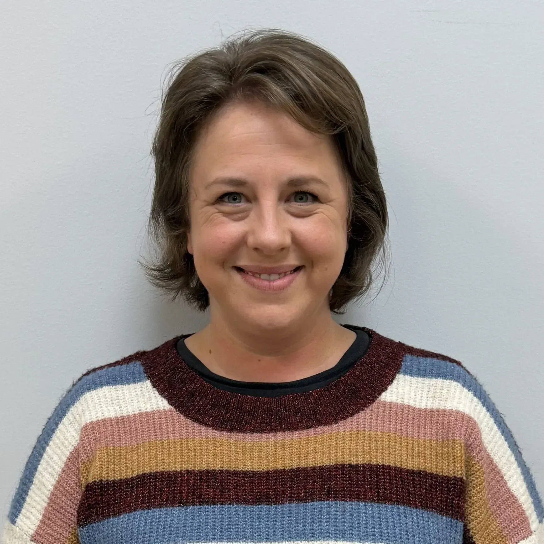 Woman smiling, wearing a striped sweater with blue, pink, and brown horizontal stripes, standing in front of a gray wall.