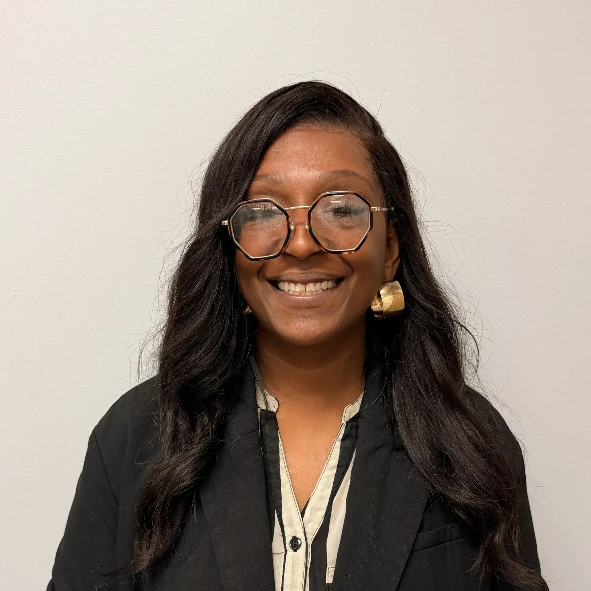 Woman with long dark hair, glasses, smiling, wearing a black blazer and gold earrings.