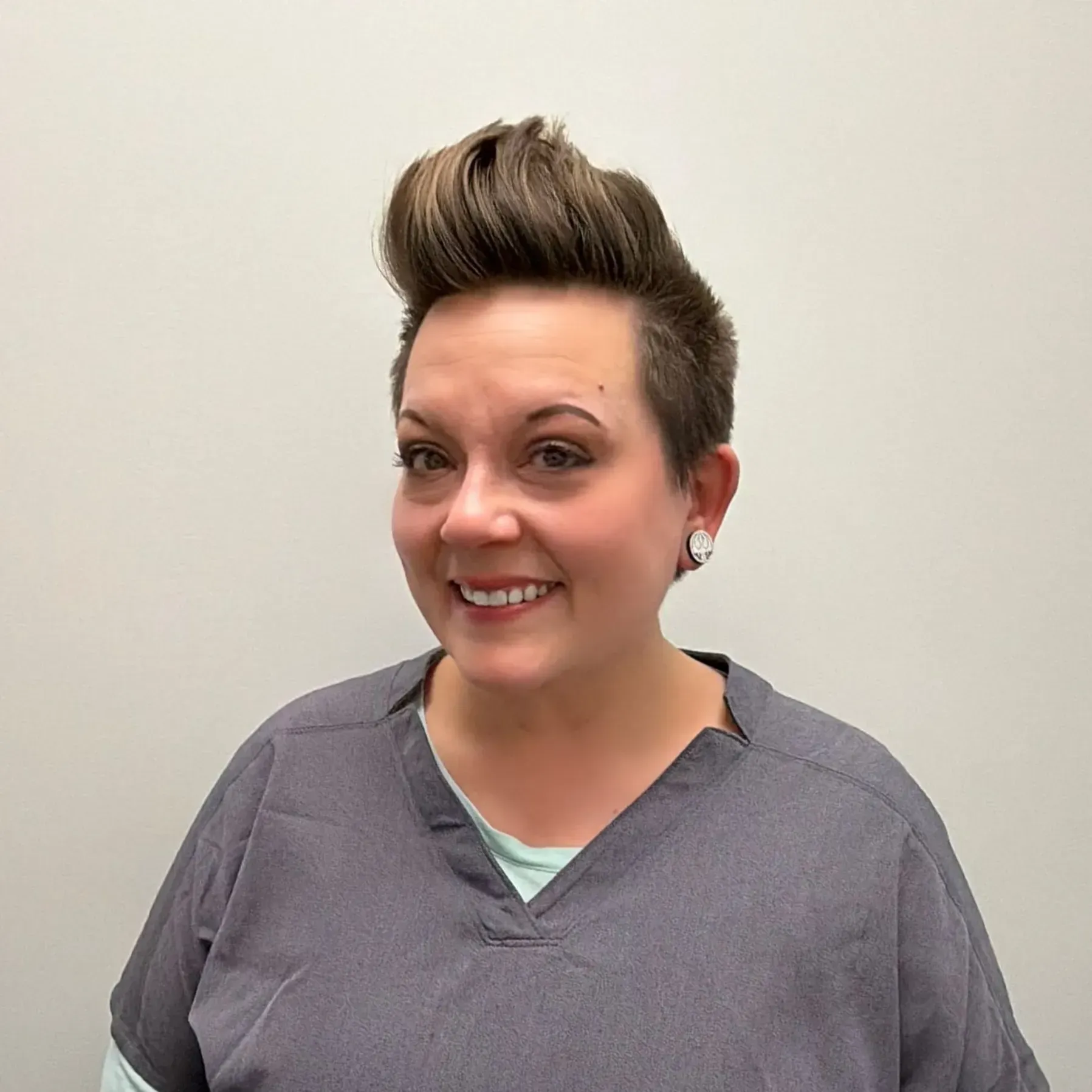 Woman with short, styled brown hair, smiling, wearing gray scrubs.