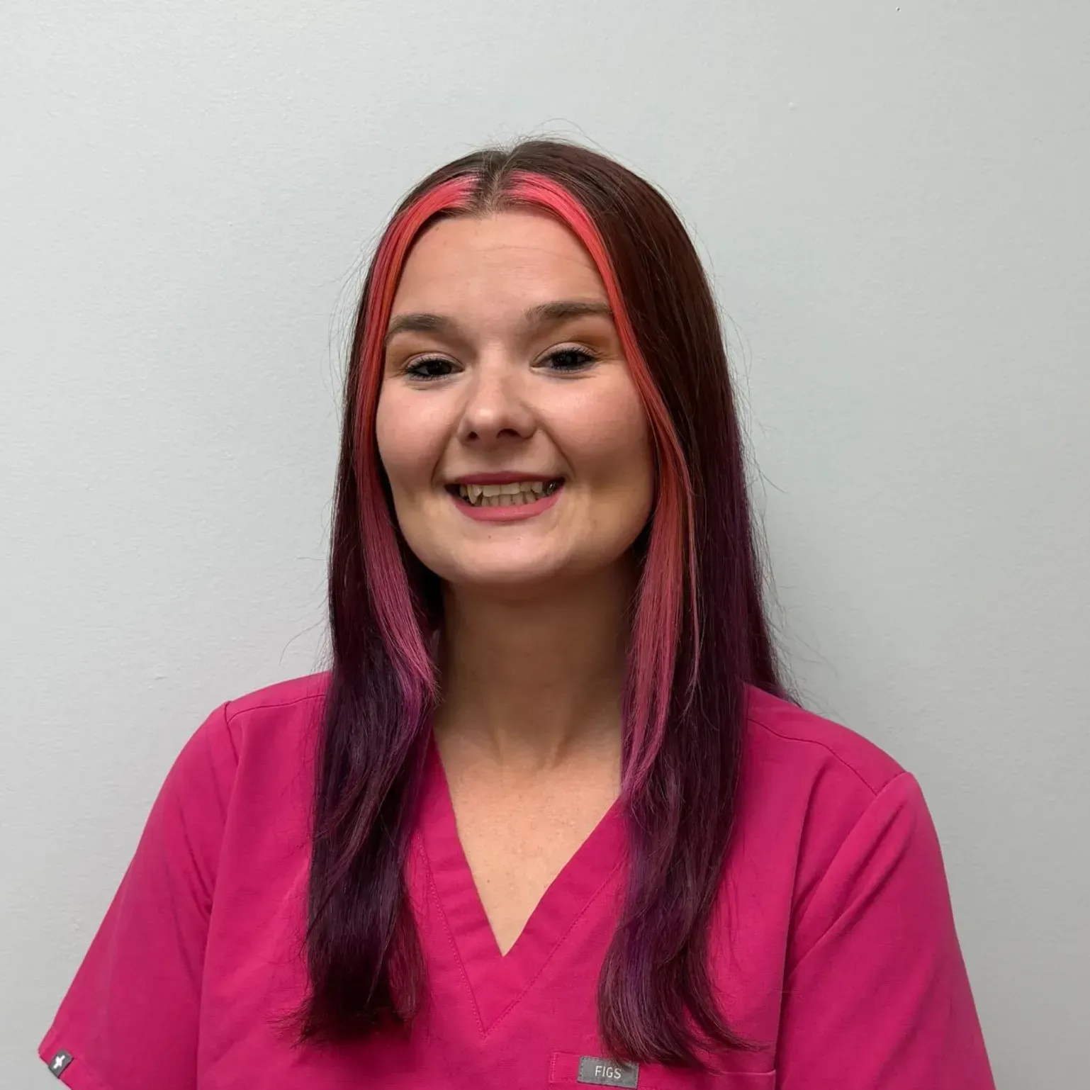 Woman with pink and purple hair, wearing a pink shirt, smiling in front of a white wall.