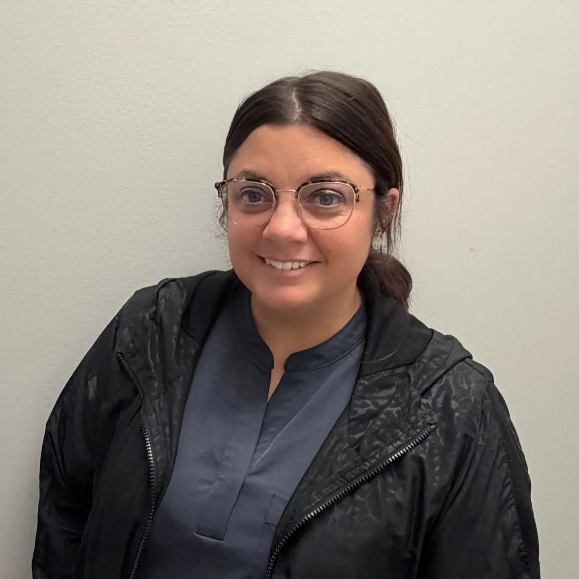 Woman with glasses smiling, wearing a dark jacket over a blue shirt, against a white wall.
