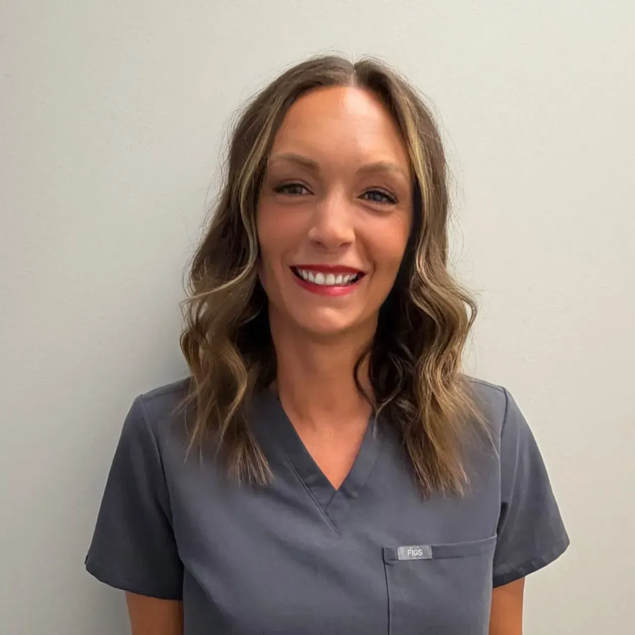 Woman with brown hair in gray scrubs smiles in a neutral setting.