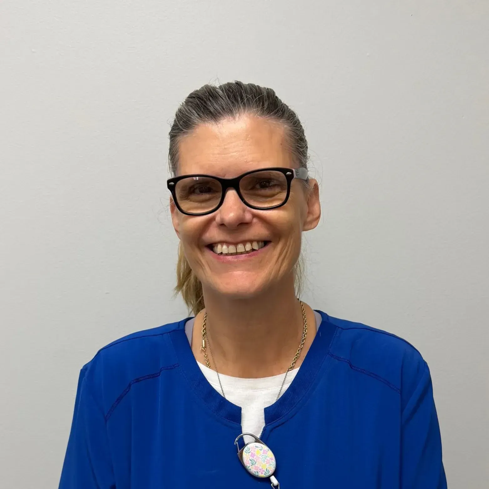 Woman with glasses and a ponytail wearing a blue scrub top, smiling.