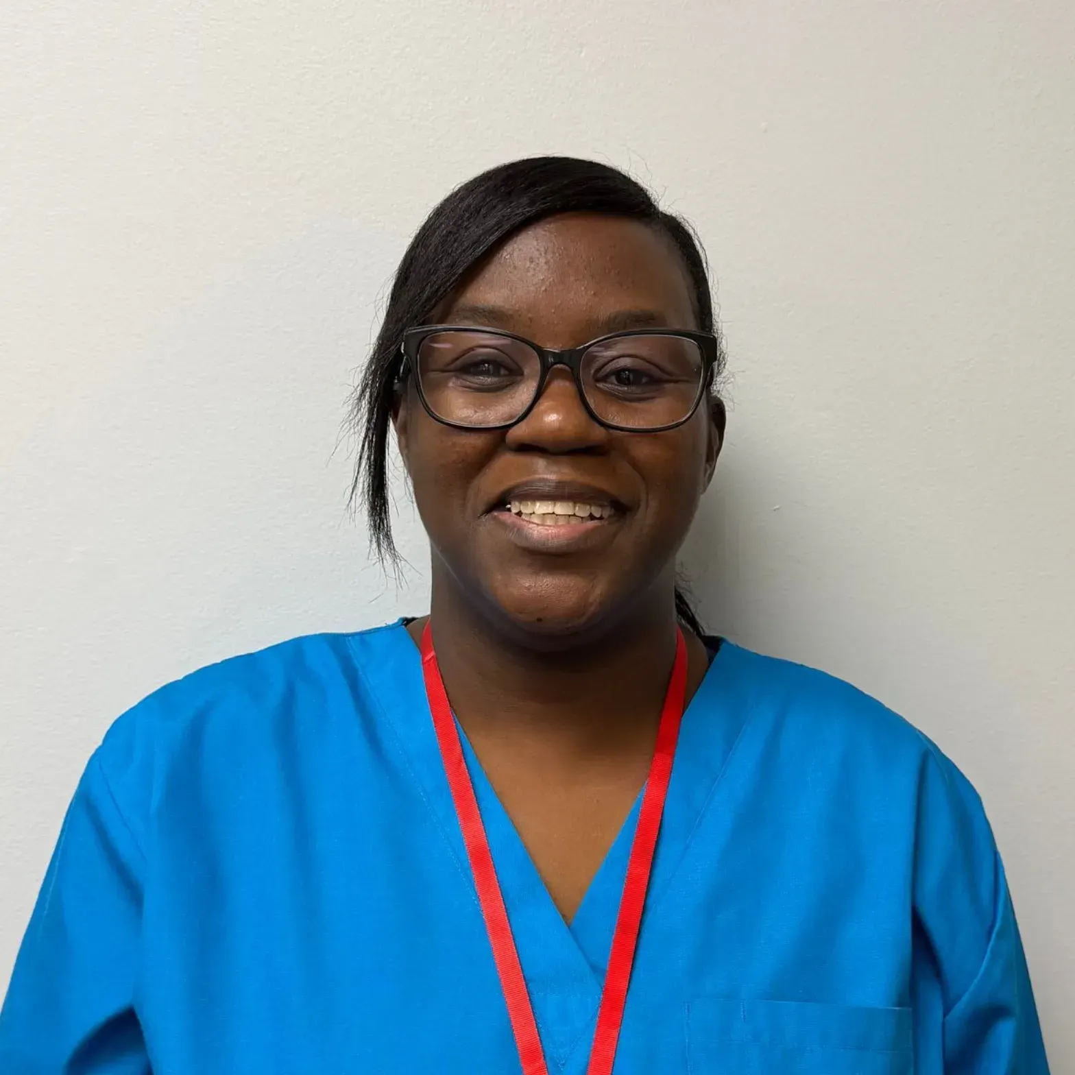 Smiling person wearing glasses and blue scrubs with a red lanyard, standing in front of a white wall.