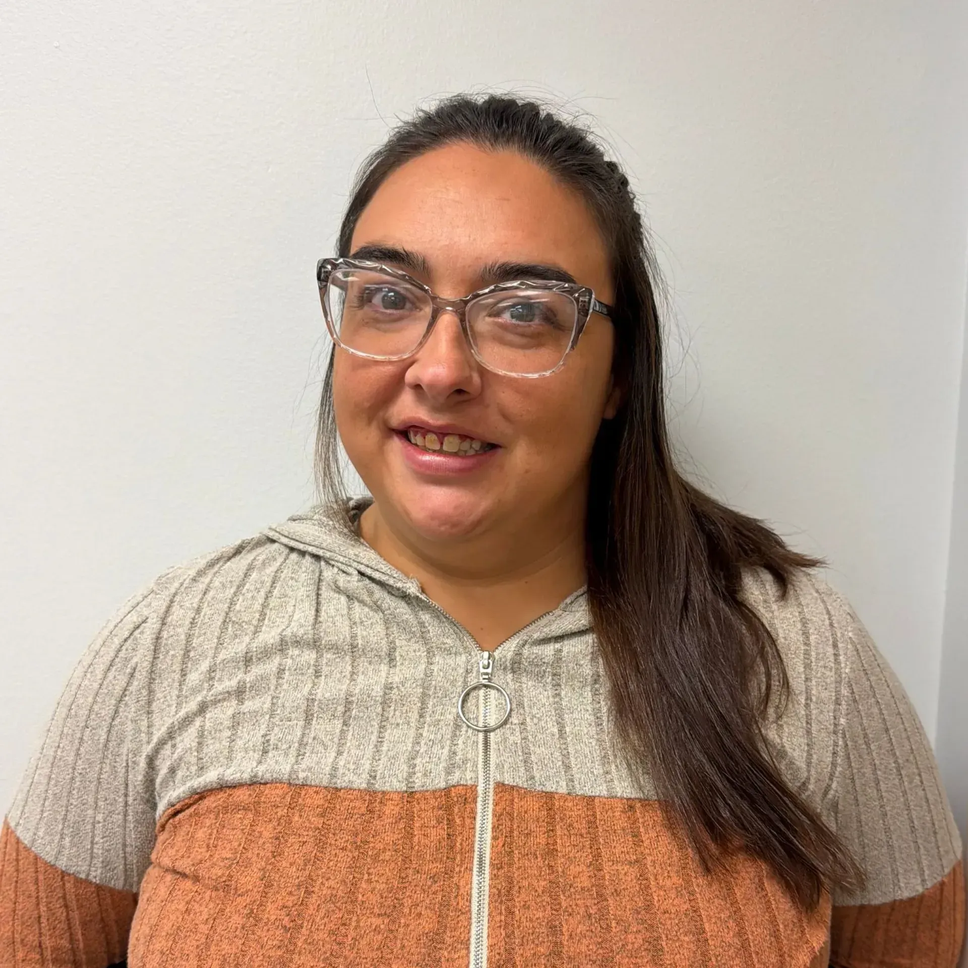 Woman with glasses and a zip-up sweater smiles in front of a white wall.