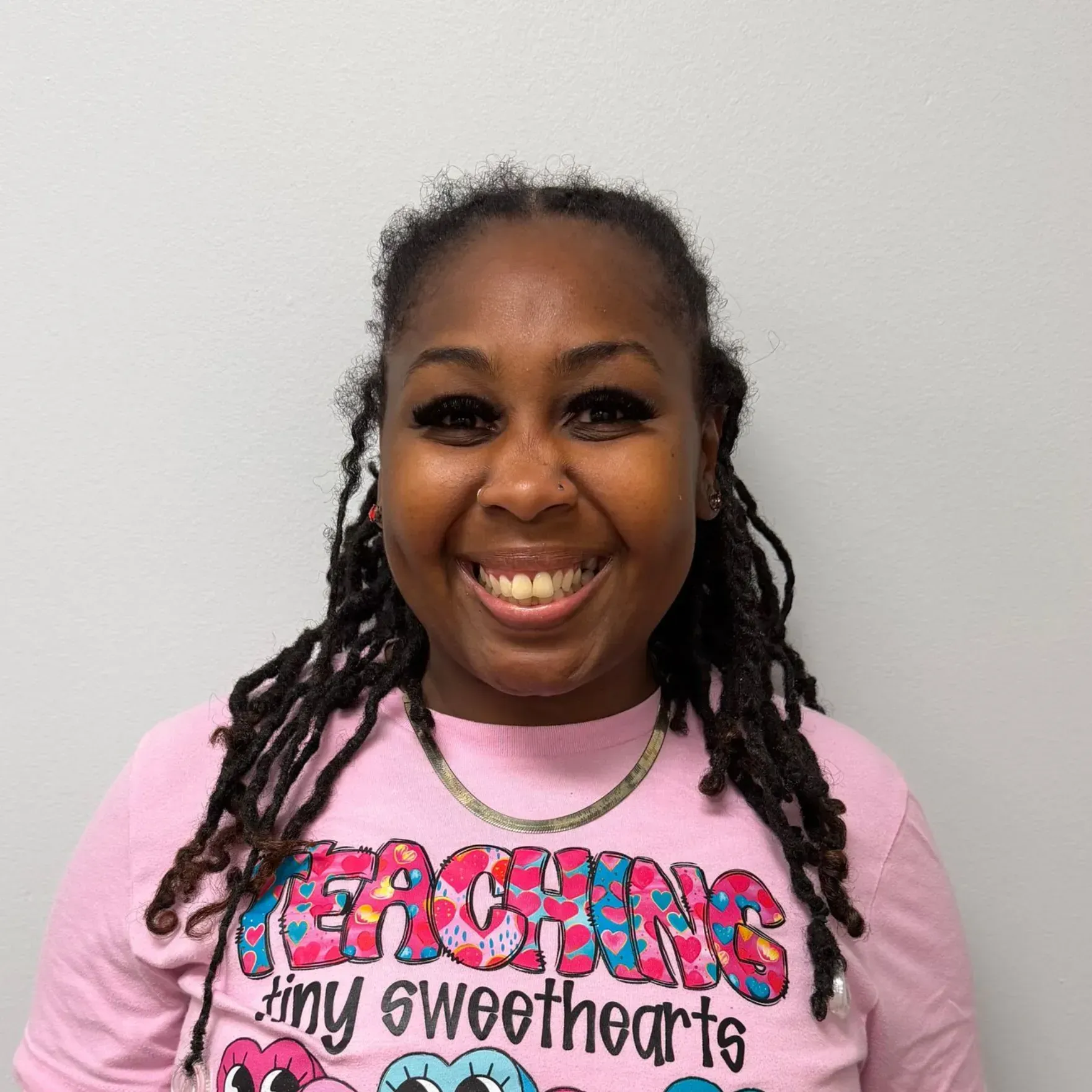 Woman with dreadlocks smiles broadly, wearing a pink shirt that says, 