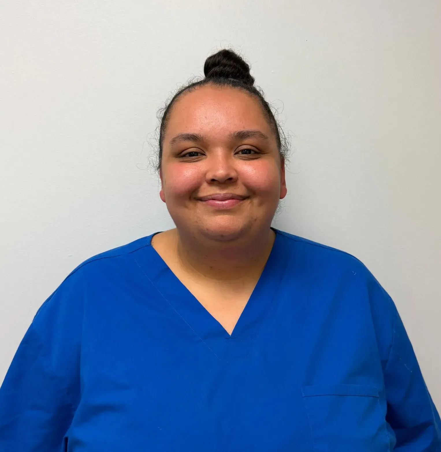 Person in blue scrubs smiles at the camera, against a white wall. Hair is up in a bun.