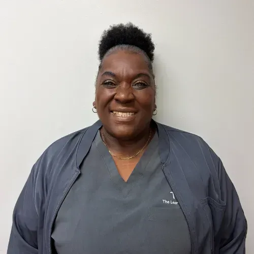Woman wearing a gray uniform smiling, standing in front of a white wall.