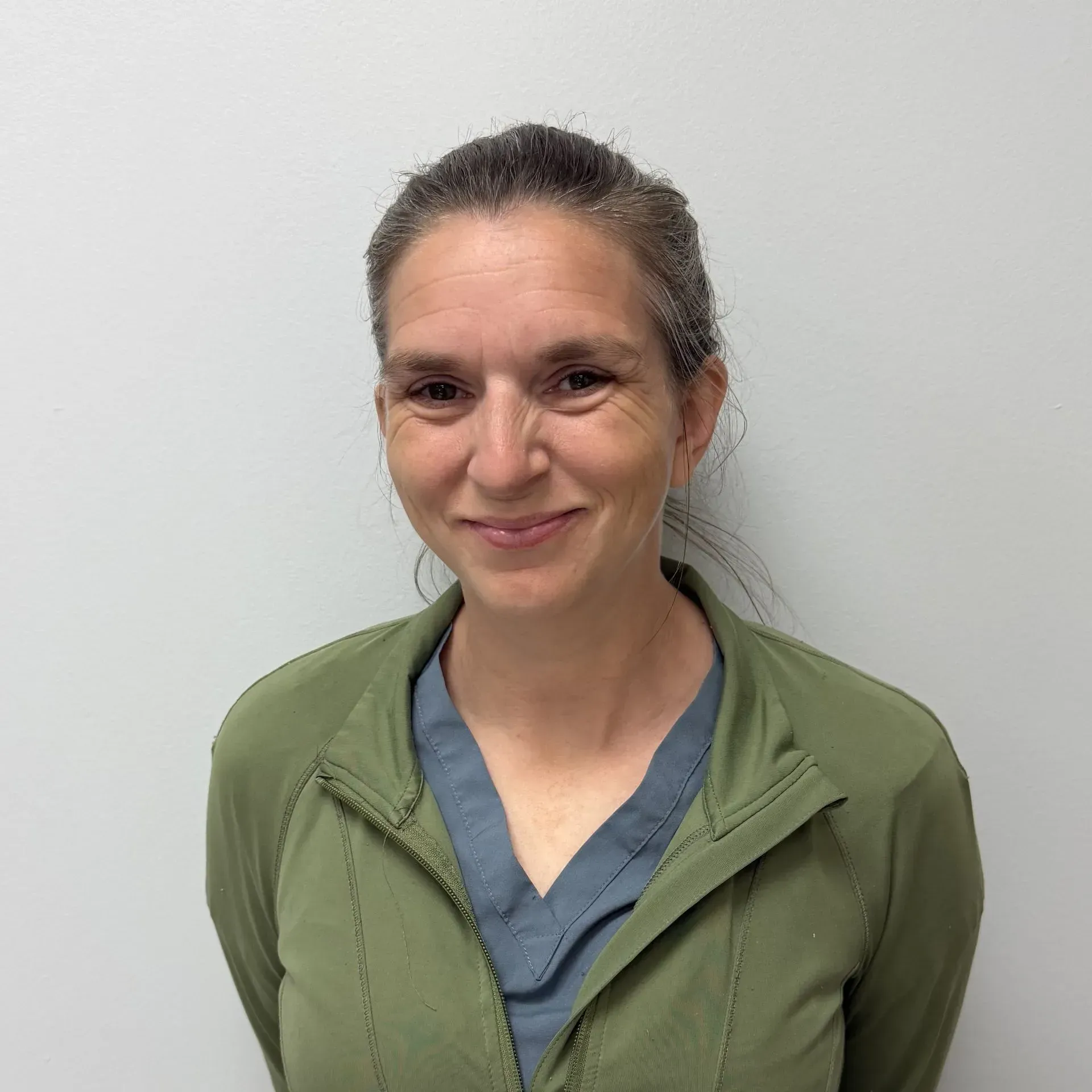 Woman in a green jacket and blue top smiles, standing against a white wall.