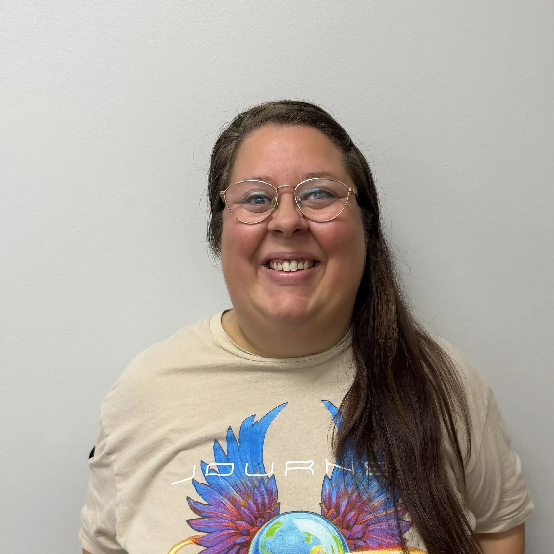 Person wearing glasses and a tan Journey t-shirt smiles in front of a gray wall.