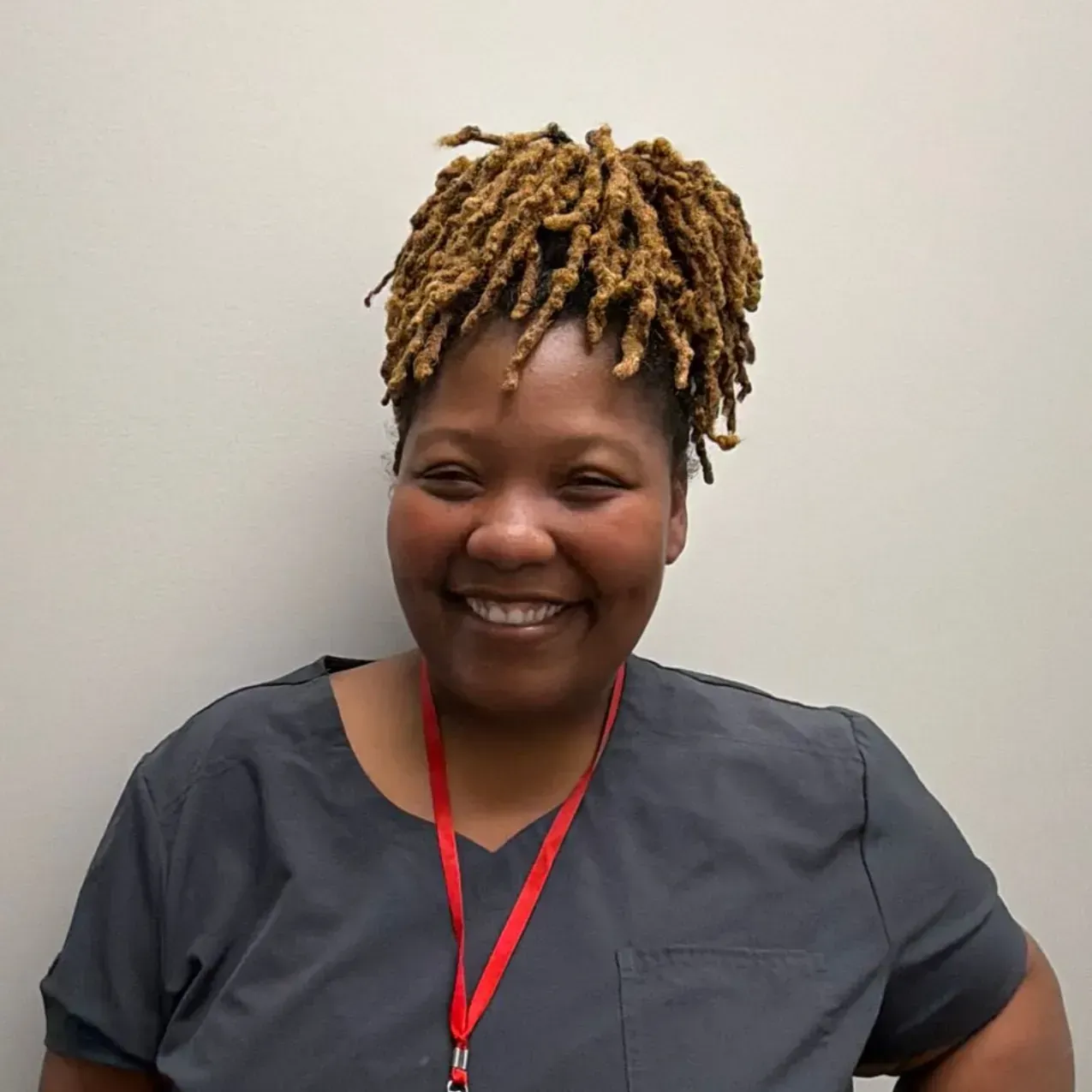 Person smiling, wearing gray scrubs and a red lanyard, against a plain wall. They have dreadlocks.