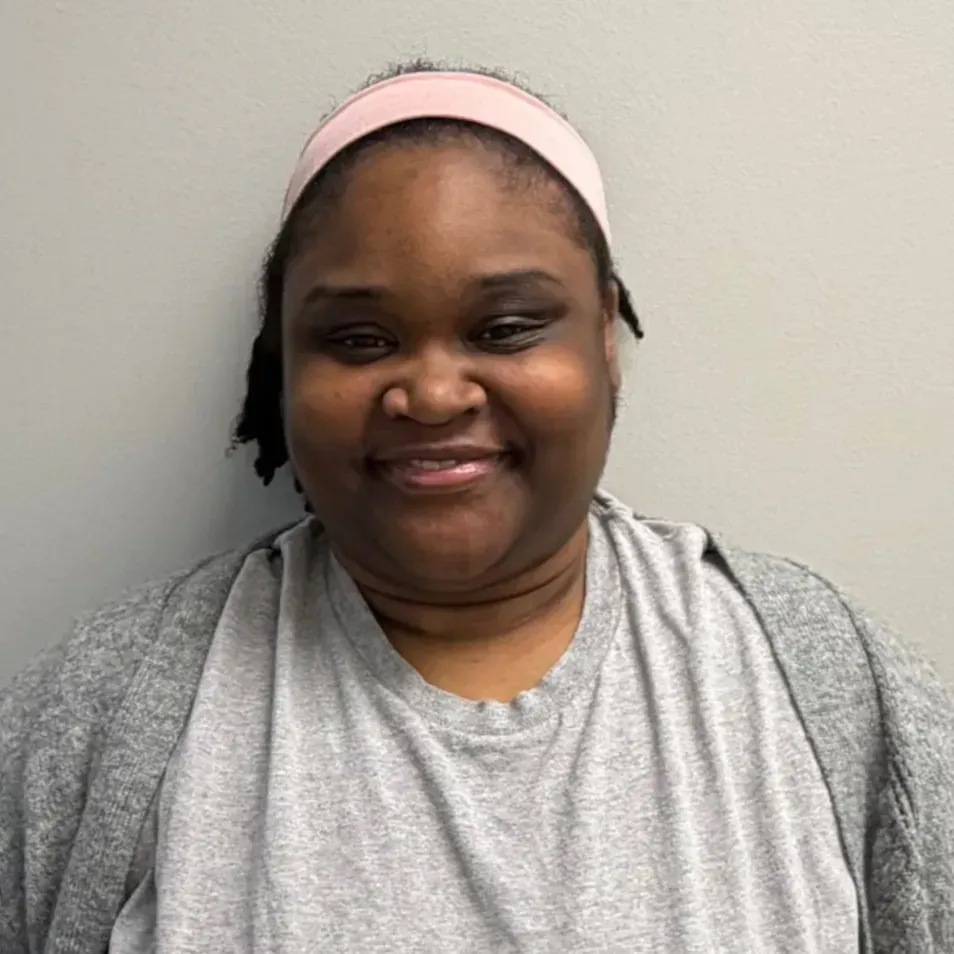 Person wearing a pink headband and gray shirt smiling against a gray wall.