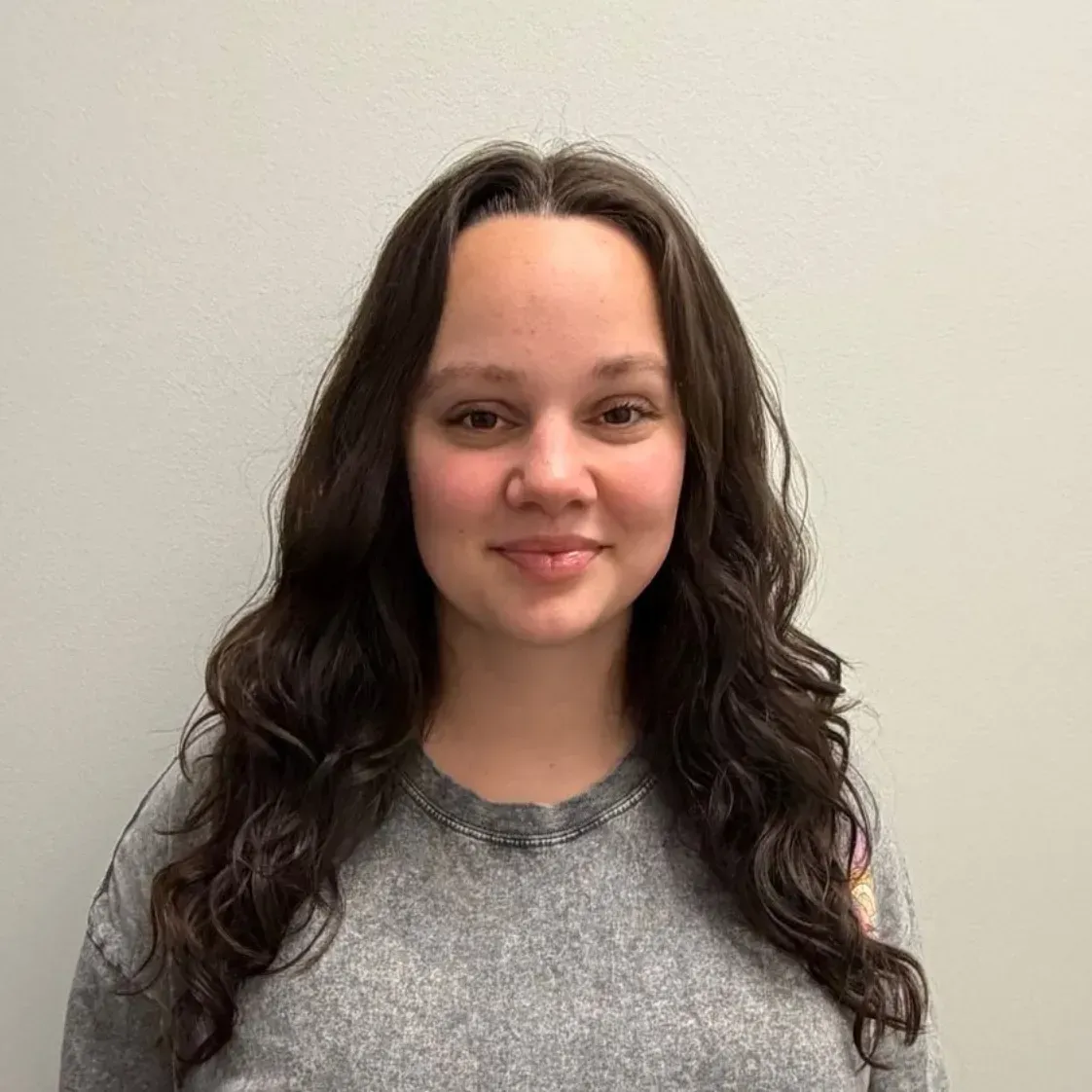 Woman with long, wavy brown hair wearing a gray sweatshirt, smiling in front of a gray wall.