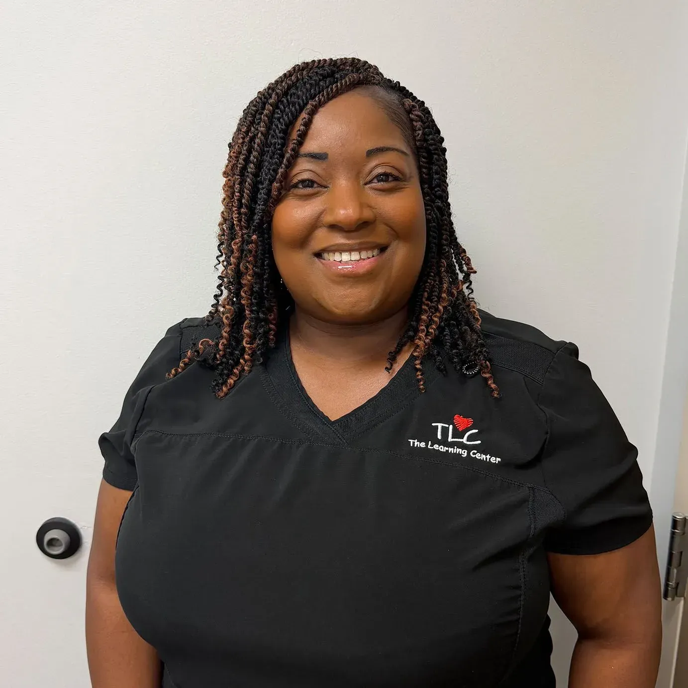 Woman in black scrubs, smiling, with braided hair, standing in front of a white wall.