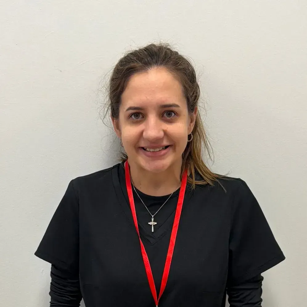 Woman wearing a black top with a red lanyard, smiling at the camera, standing in front of a white wall.