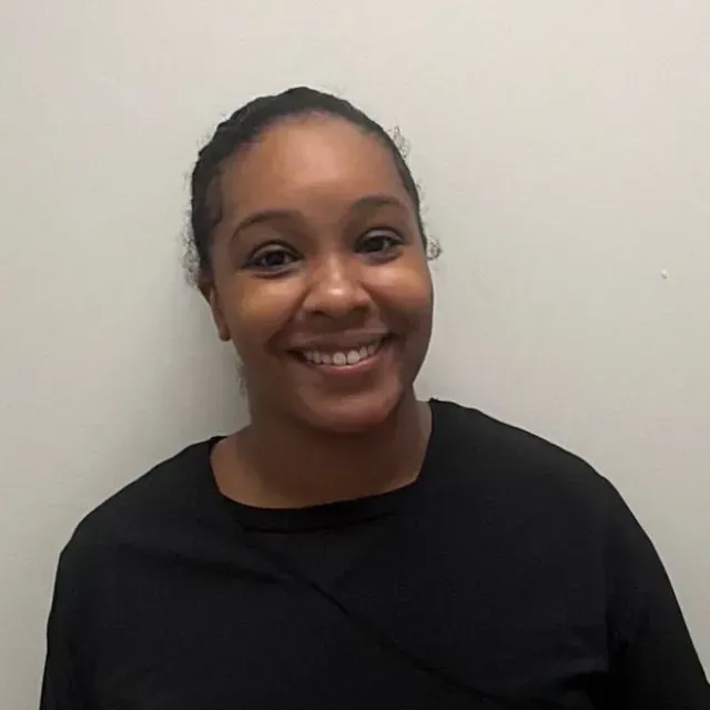 Woman in black top smiling, standing against a white wall.