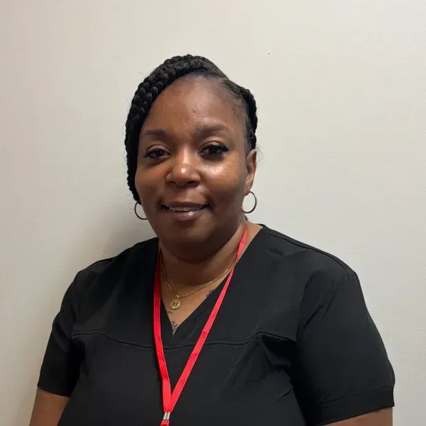 Woman with braided hair, wearing black scrubs and a red lanyard, smiling in front of a white wall.