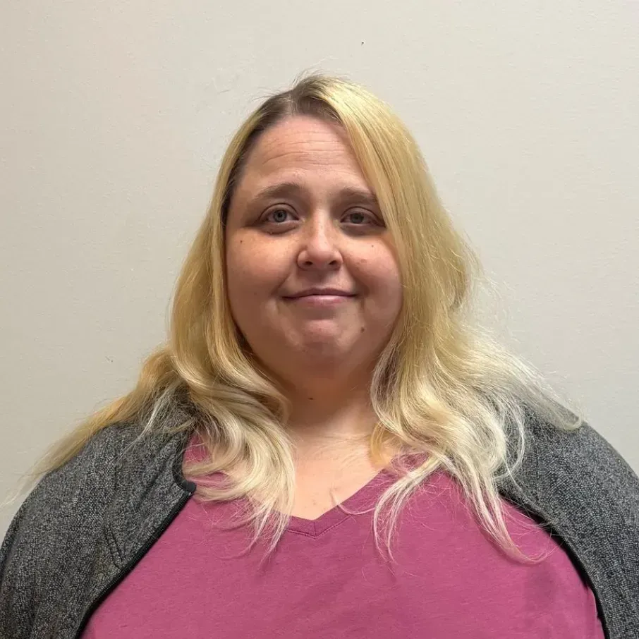 Woman with long blonde hair wearing a pink shirt and grey cardigan smiles at the camera against a neutral background.