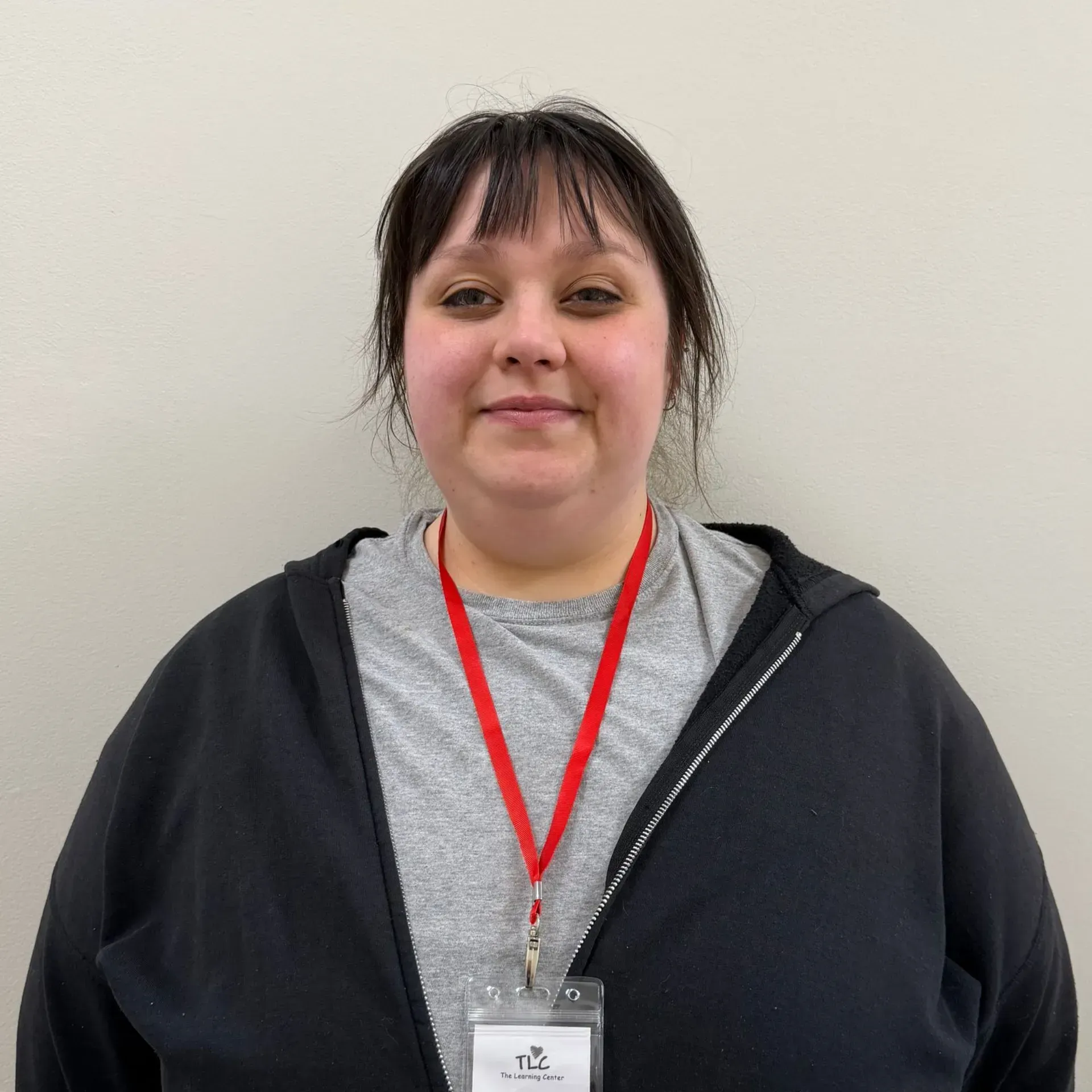 Woman with dark hair, wearing a black jacket and gray shirt, red lanyard with an ID badge, smiling in front of a gray wall.