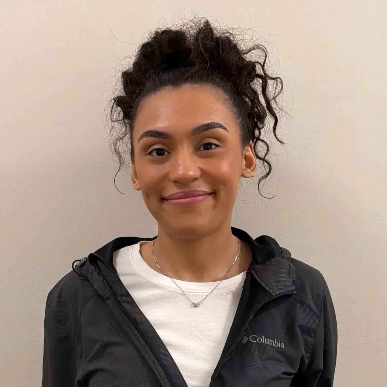 Woman with dark curly hair in a bun smiling, wearing a white shirt and black Columbia jacket.