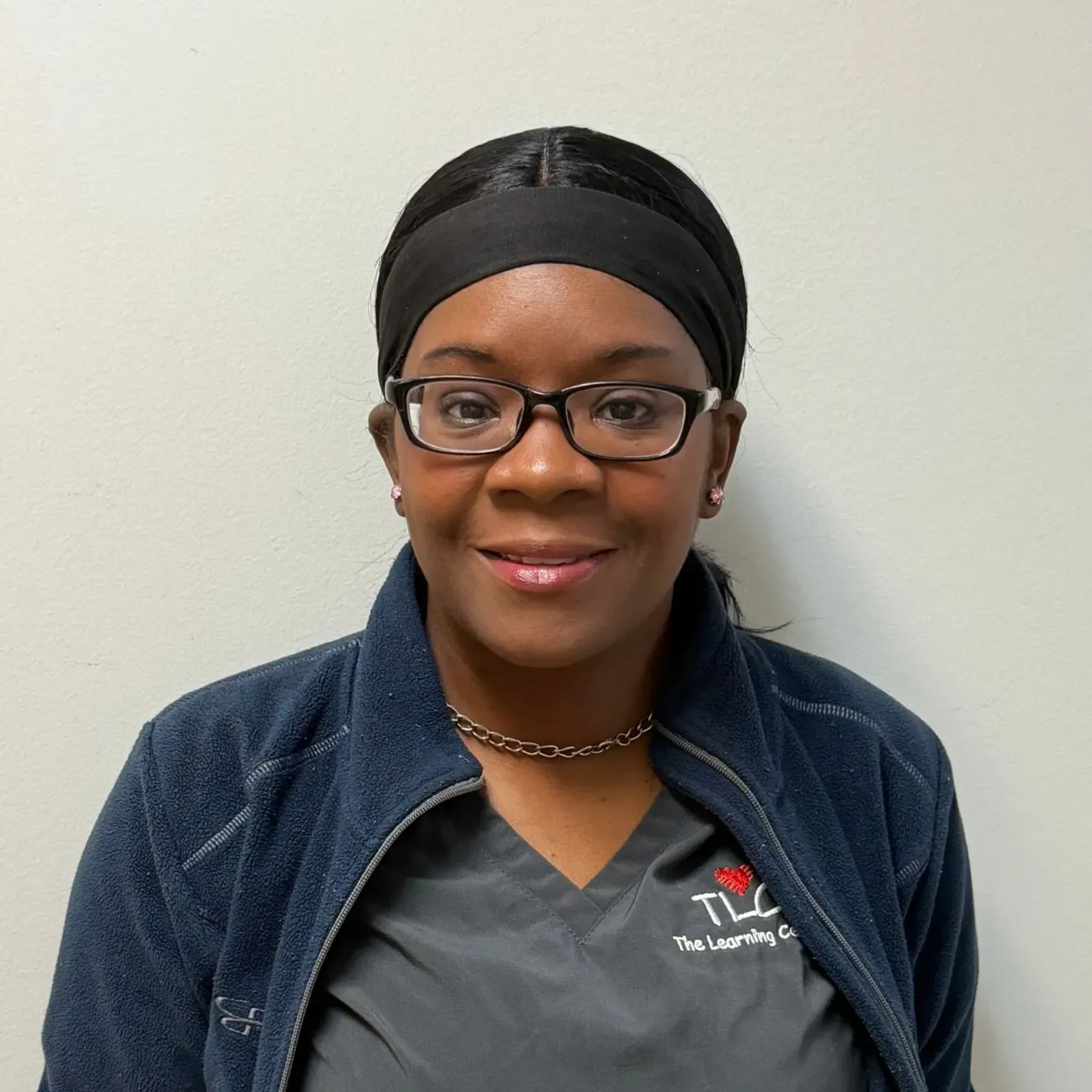 Woman wearing glasses, a black headband, and a gray TLC Learning Center uniform smiling.