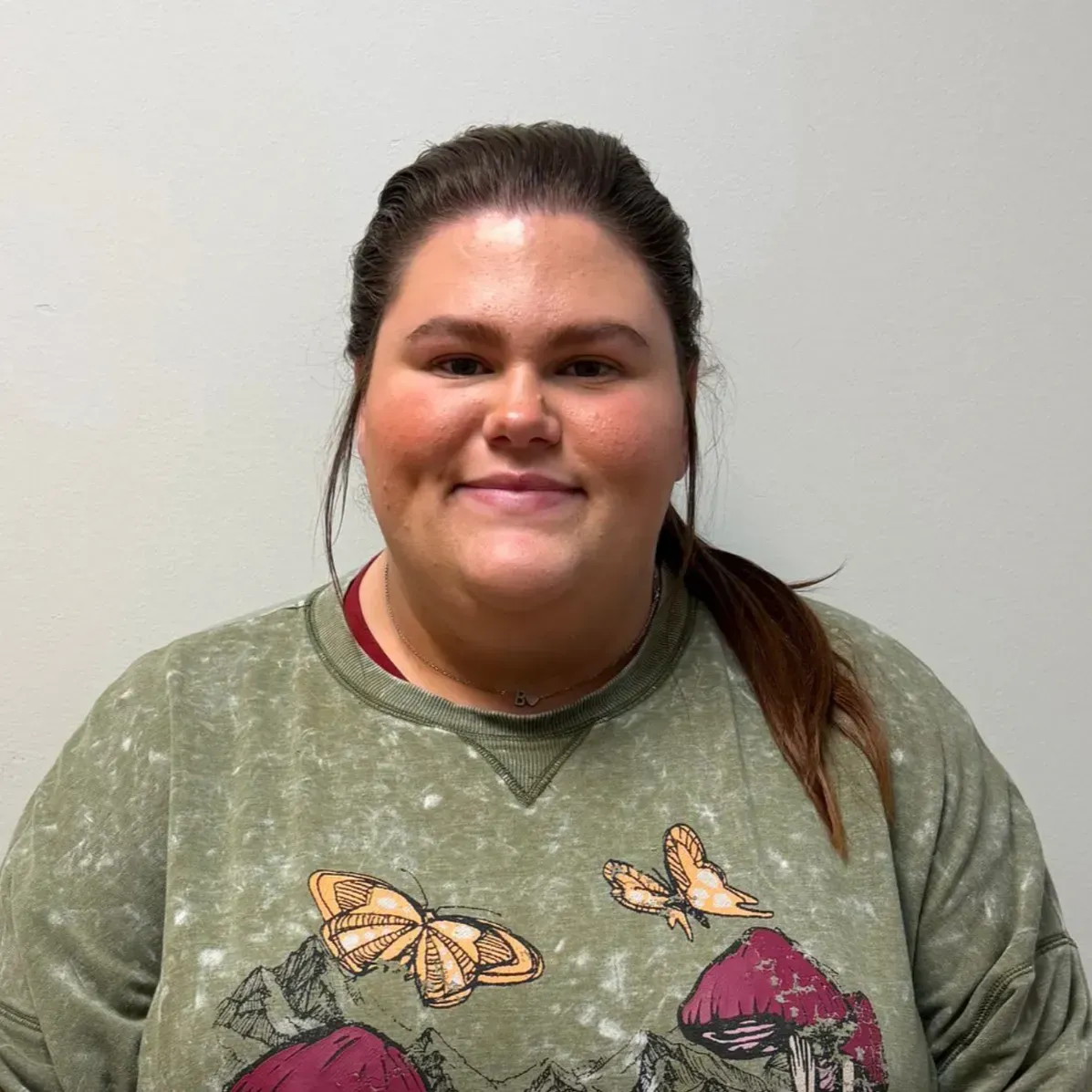 Woman with dark hair in a ponytail, wearing a green sweatshirt with butterfly design, smiles at the camera.