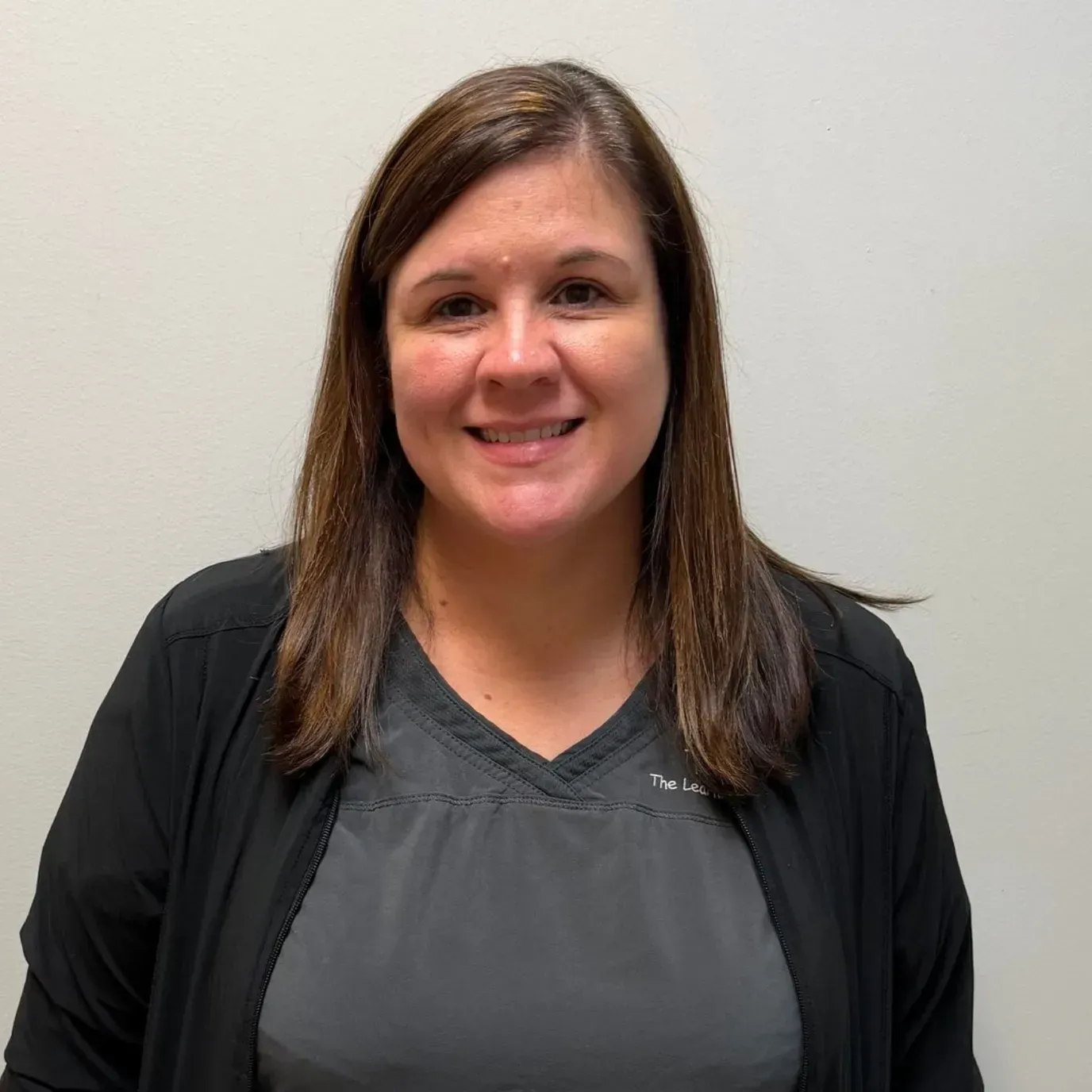 Woman with brown hair smiles, wearing a black jacket and gray top in front of a white wall.