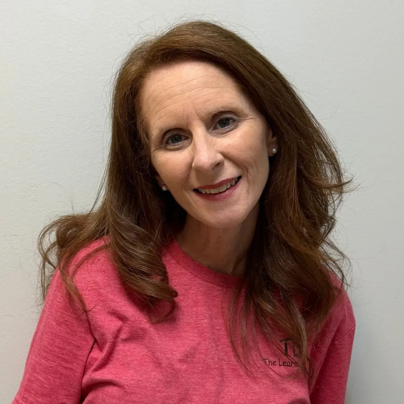Woman with reddish-brown hair smiles, wearing a pink shirt, in front of a white wall.