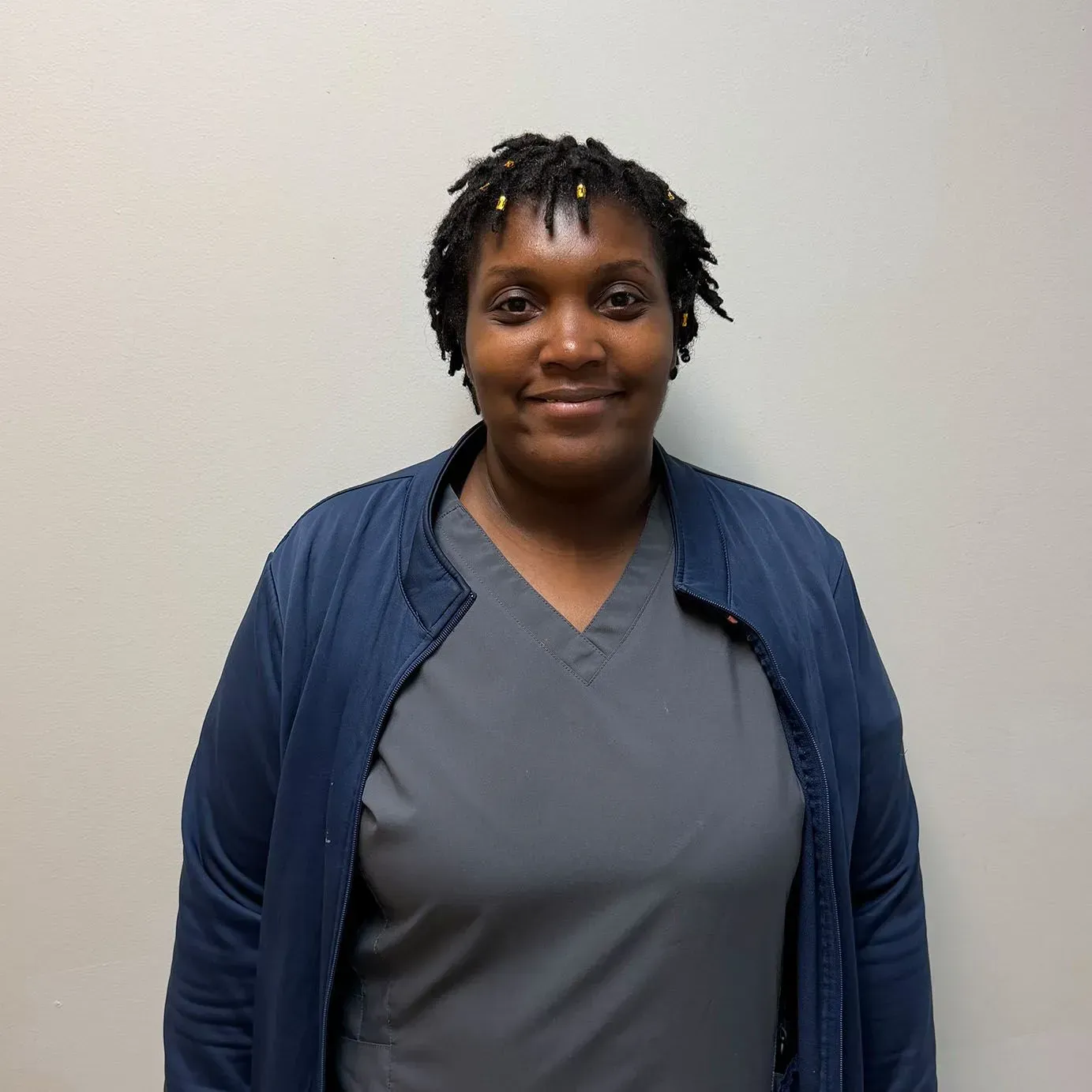 Woman in blue jacket and grey top against a light wall. Smiling.