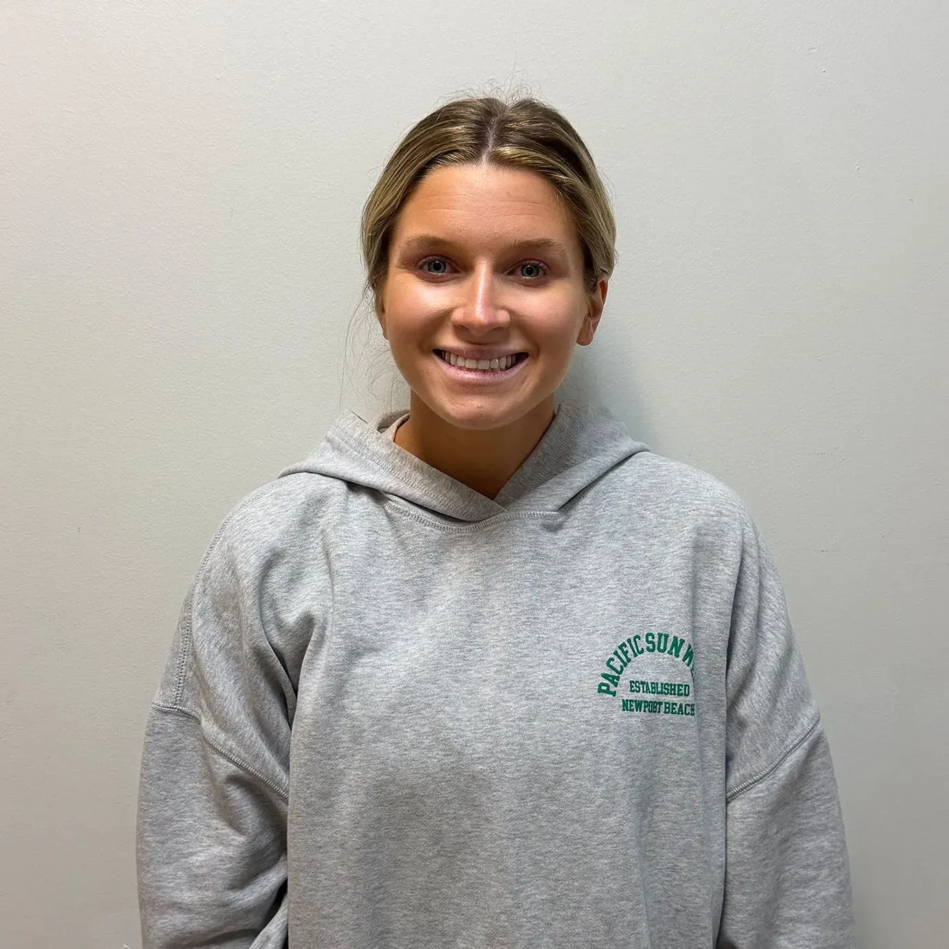 Woman in gray hoodie smiles, standing against a neutral-colored wall.