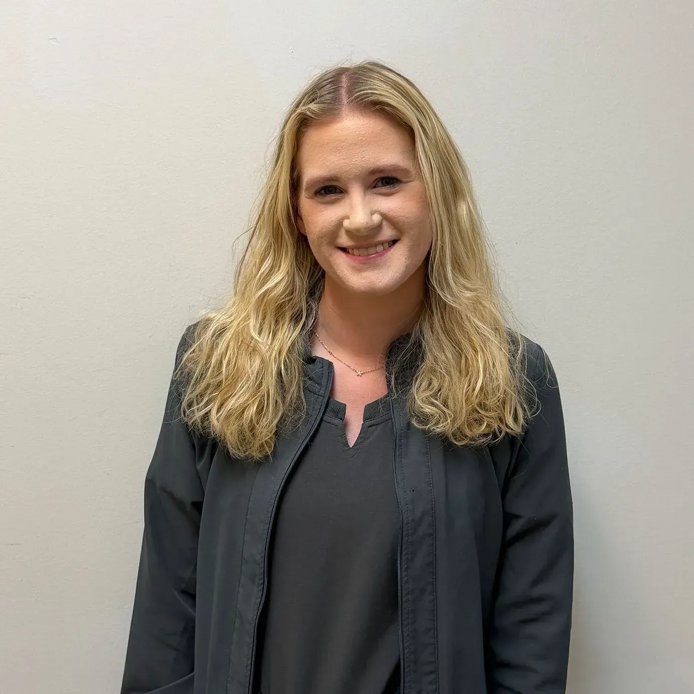 Woman with blonde hair wearing a grey jacket and shirt smiles in front of a neutral wall.