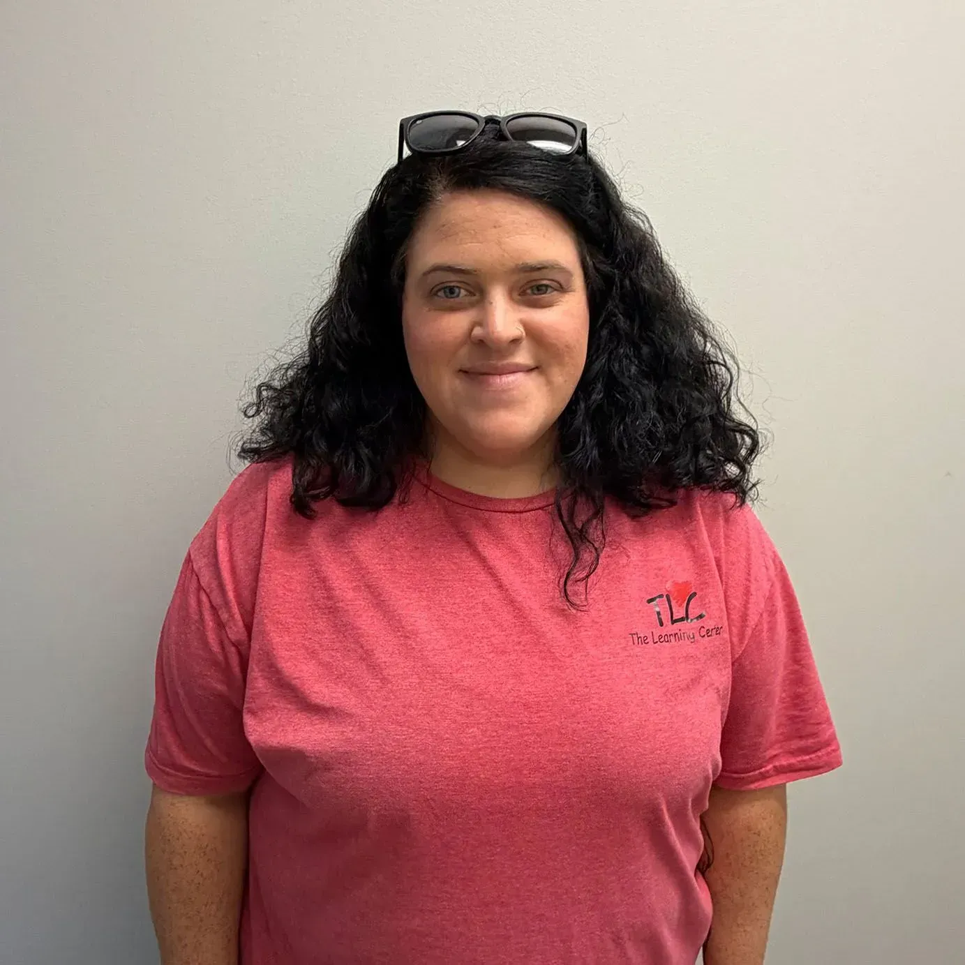 Woman with dark curly hair wearing a red t-shirt, glasses on her head, smiles at the camera.