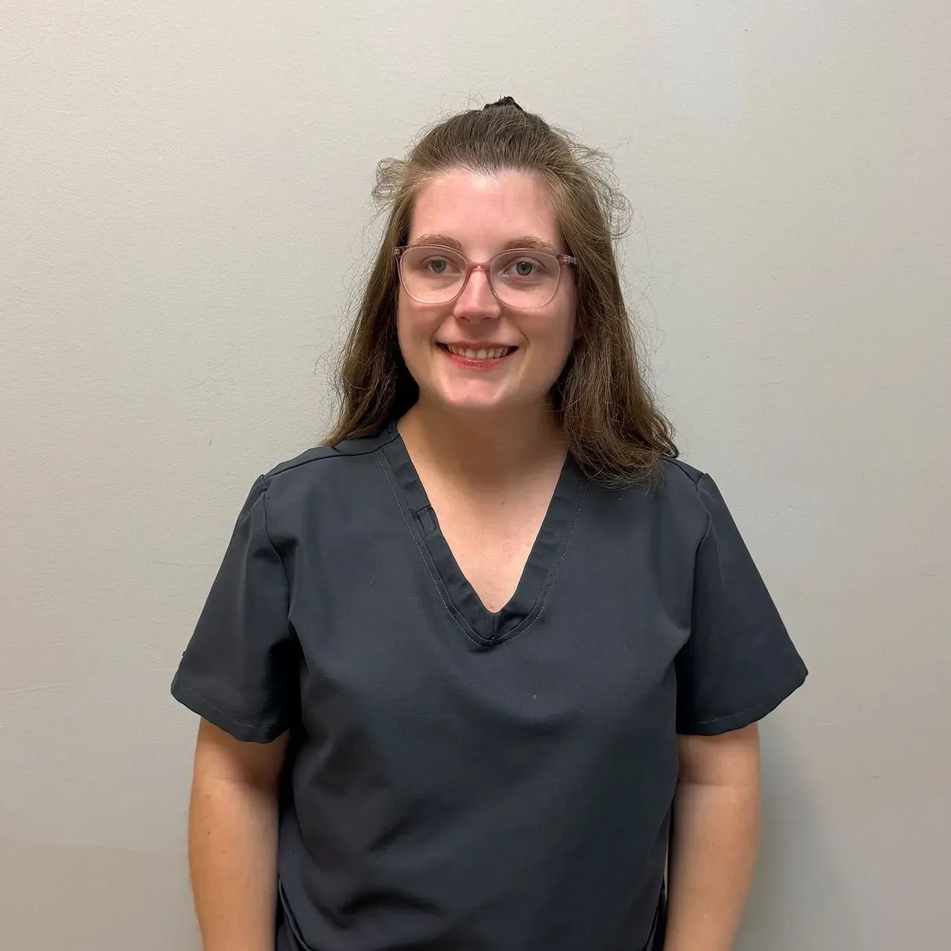 Woman with glasses in gray scrubs smiles in front of a light wall.