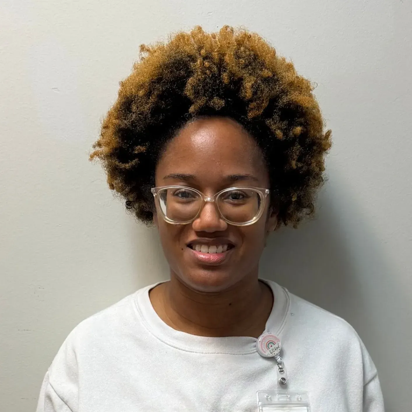 Woman with curly brown hair, glasses, and a name tag smiles at the camera, wearing a light-colored top.