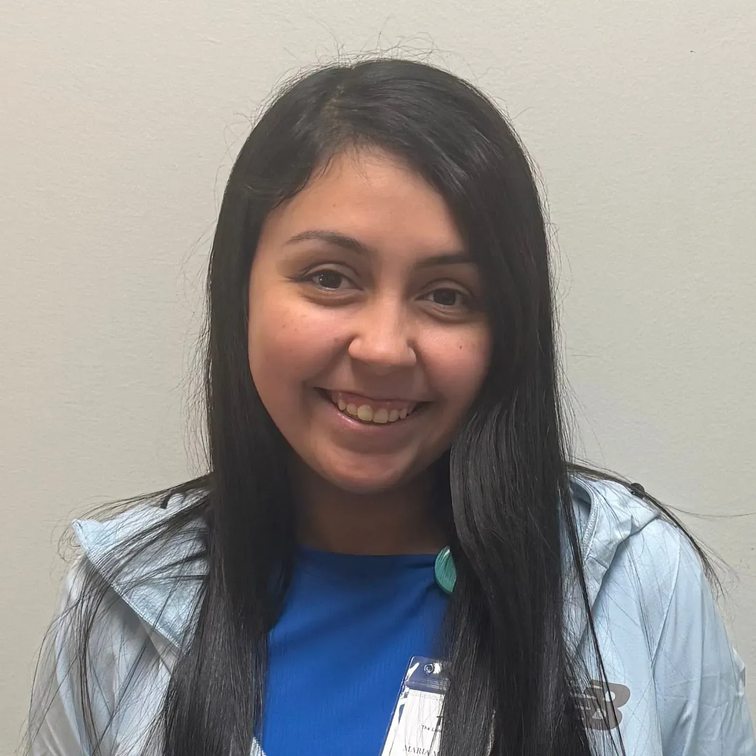 Woman with dark hair smiles, wearing a blue shirt and light blue jacket. Light-colored wall in the background.