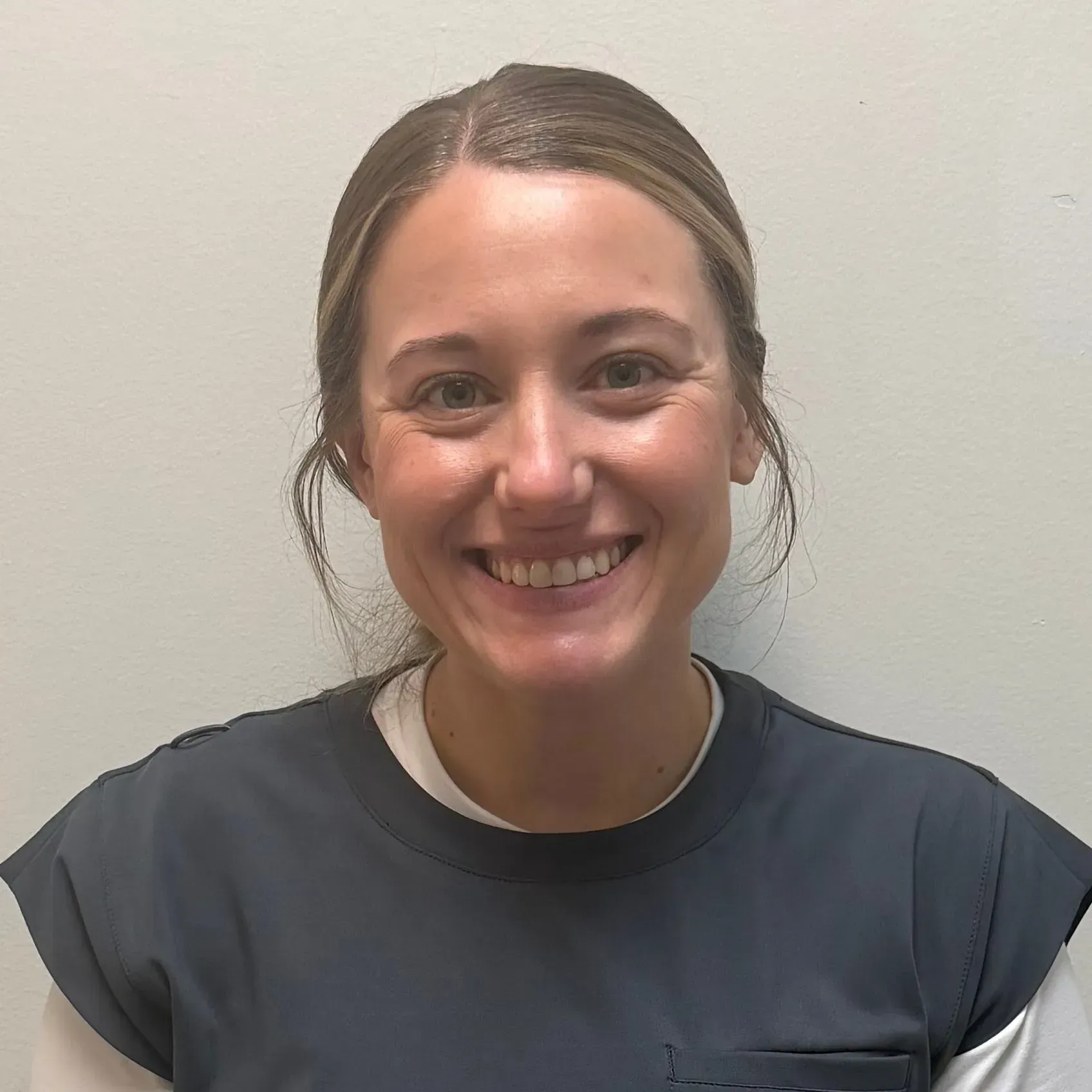 Woman smiling, wearing a gray top and white shirt, against a plain white wall.