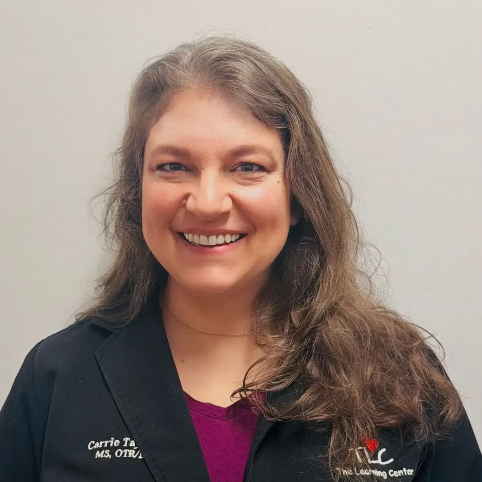 Woman with brown hair smiles, wearing a black coat with a name tag, a maroon shirt, and standing in front of a gray wall.