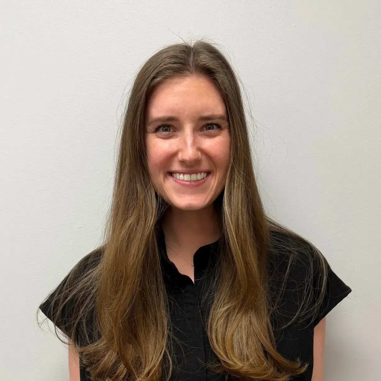 Woman with long brown hair, smiling, wearing a black top against a white background.