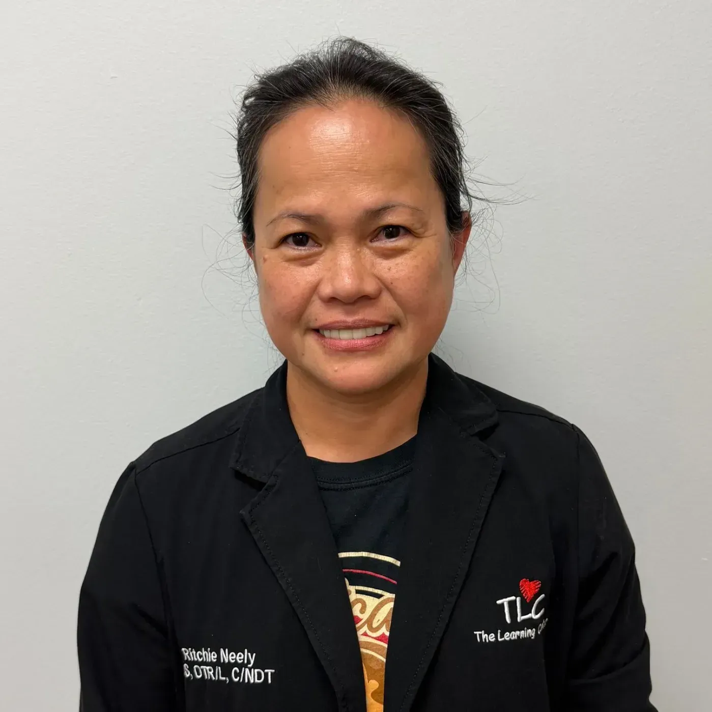 Woman wearing a black jacket with logo, smiling, standing in front of a white wall.