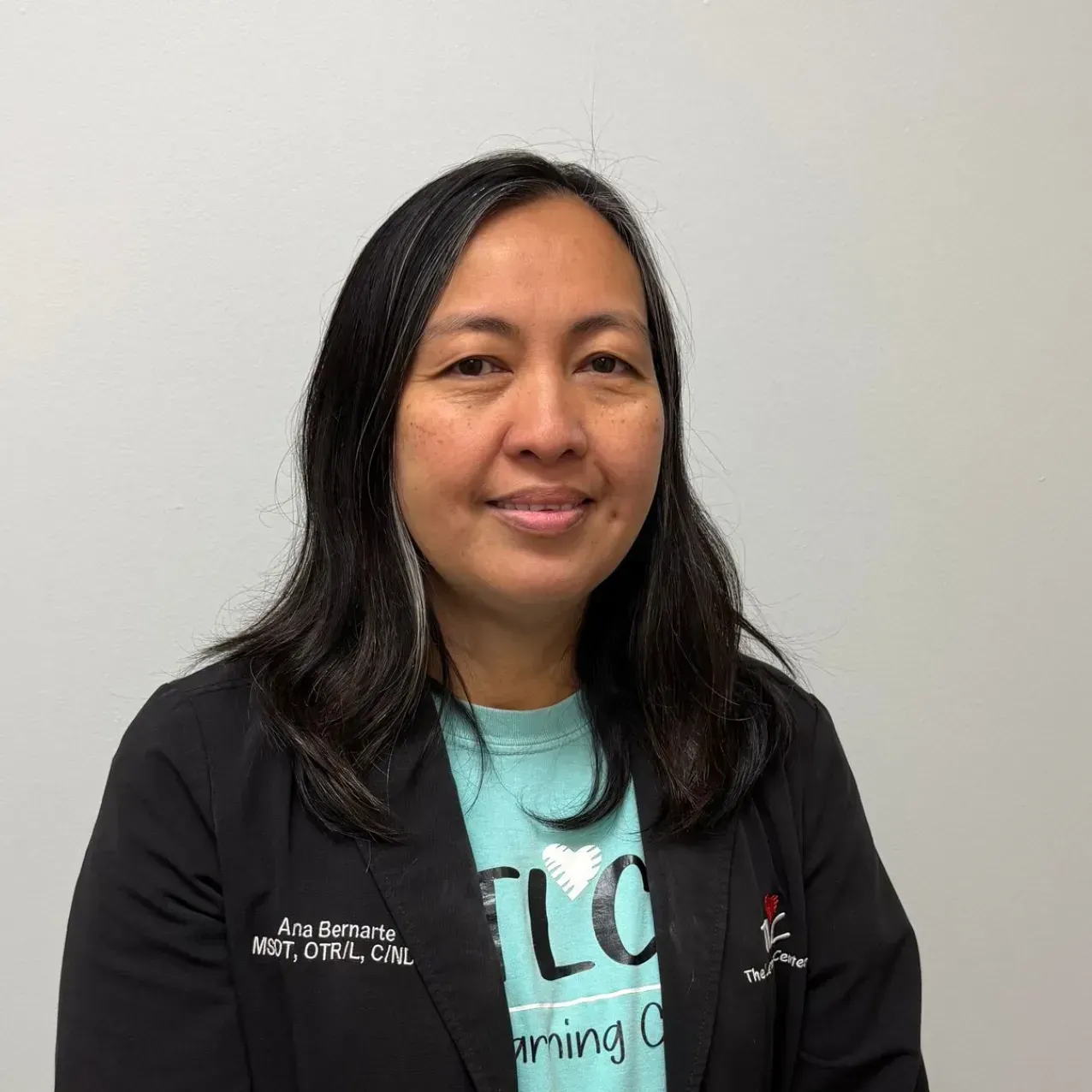 Woman with long dark hair smiles, wearing a black blazer over a turquoise shirt, in front of a white wall.