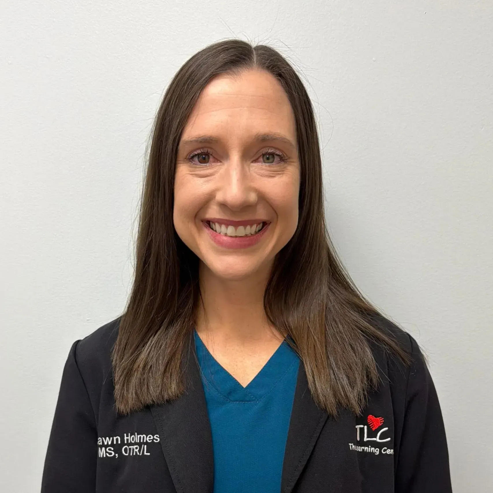 Woman with dark hair smiling, wearing a black blazer and teal scrub top. 