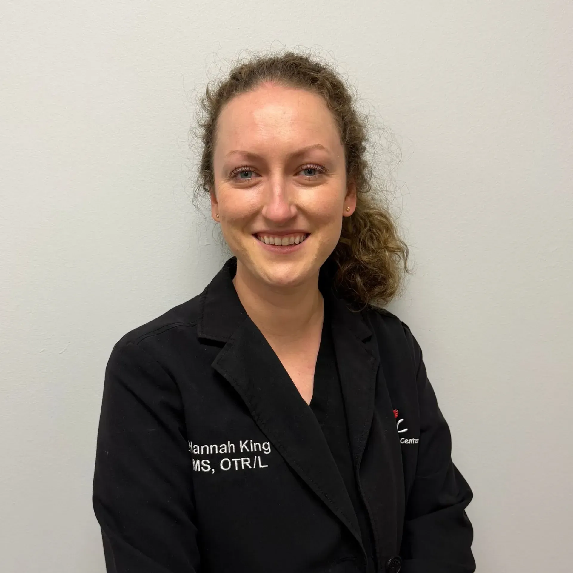 Woman with curly hair in a black blazer smiling at the camera. Text on blazer: 