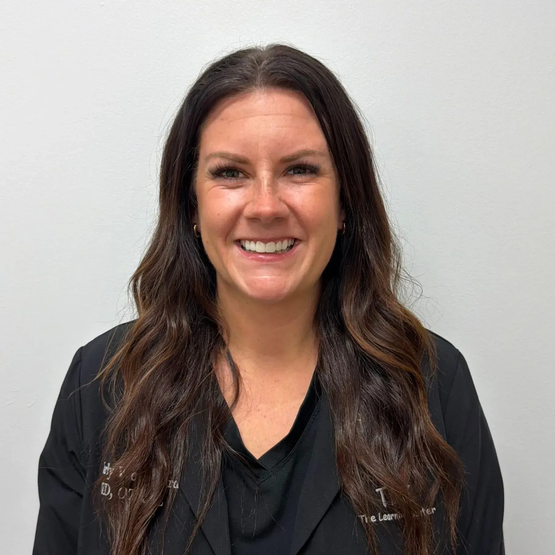 Woman with long brown hair smiles, wearing black jacket against white wall.