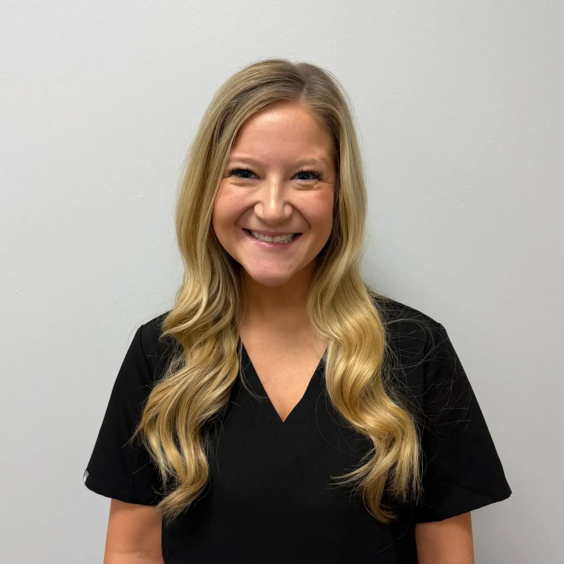 Woman with blonde hair wearing a black top, smiling in front of a gray wall.