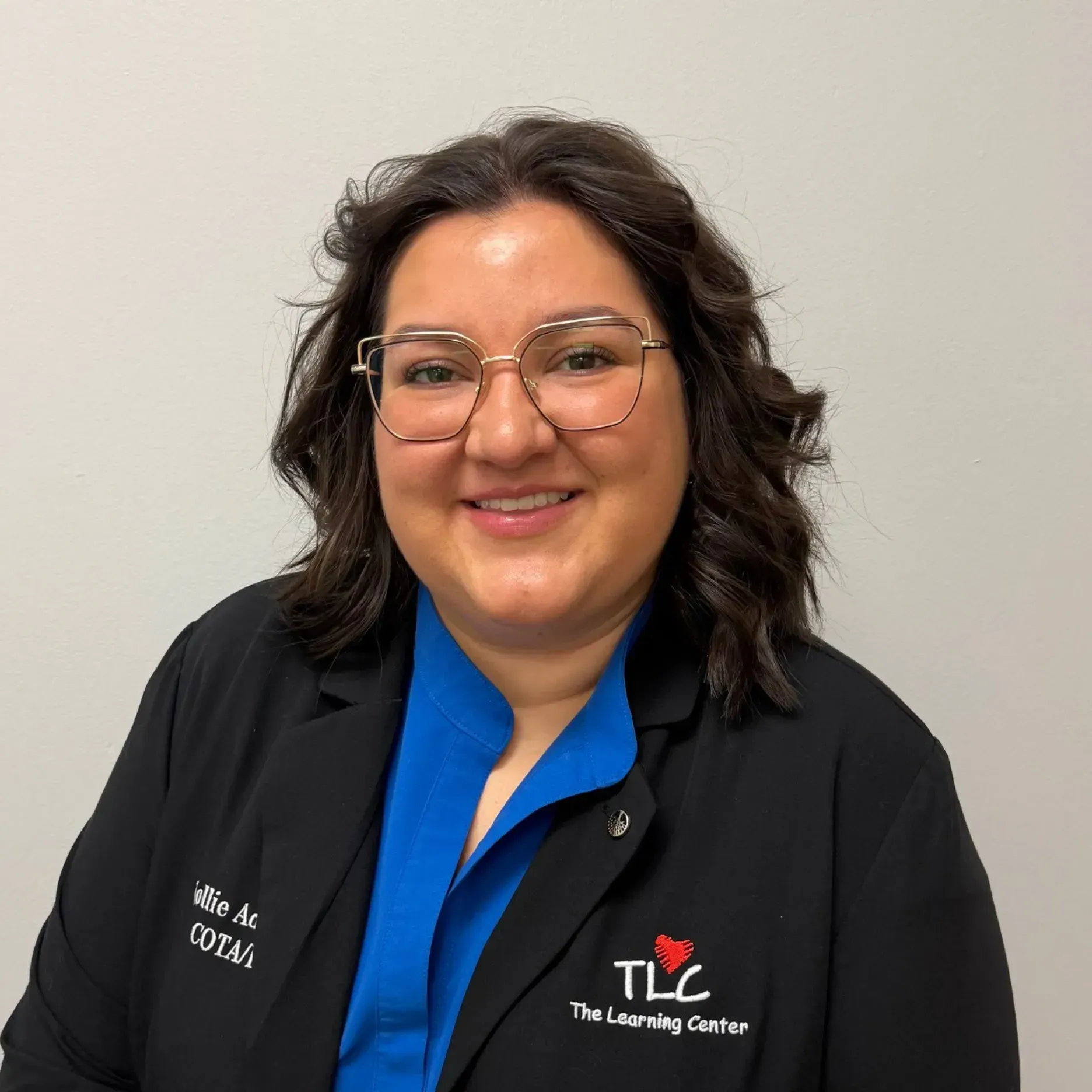 Woman with glasses smiling, wearing a blue shirt and black jacket with a 