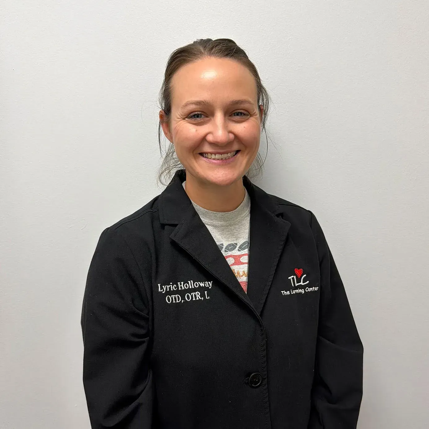 Woman in a black lab coat, smiling, standing in front of a white wall. Name tag says: Lyric Sullivan, OTD, OTR/L.